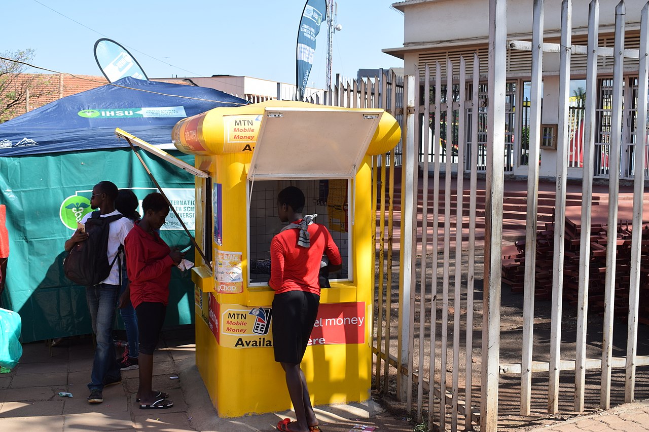 Mobile money kiosk storefront in Uganda with telecom branding and street activity.